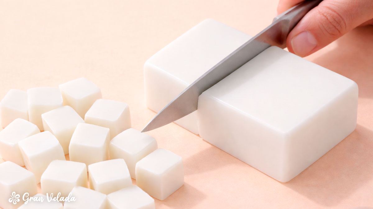 Grande plano de uma mão a cortar um bloco de base de sabonete de glicerina branca em cubos pequenos com uma faca sobre uma superfície clara.