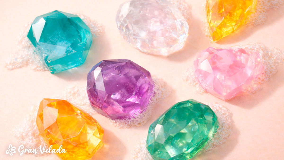 Vários sabonetes de glicerina com formato de pedras preciosas lapidadas em cores translúcidas como turquesa, ametista e âmbar, rodeados por espuma branca.