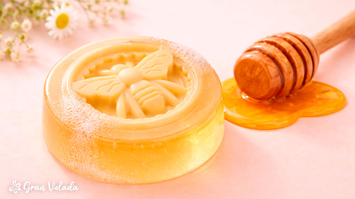 Sabonete de glicerina circular de cor âmbar translúcida com o relevo de uma abelha no centro, acompanhado por uma colher de pau com mel real.