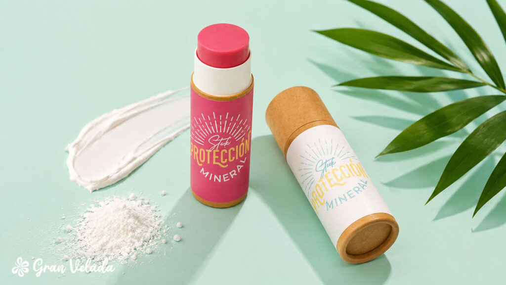 Duas embalagens de cartão tipo stick de protetor solar mineral sobre um fundo verde-claro, acompanhadas por uma amostra de creme branco, pó mineral e uma folha de palmeira.