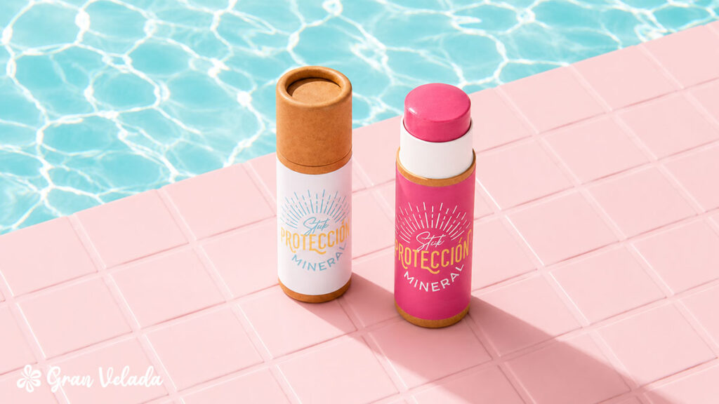 Dois recipientes em barra de protetor solar mineral colocados junto à borda de uma piscina com água cristalina e azulejos cor-de-rosa.